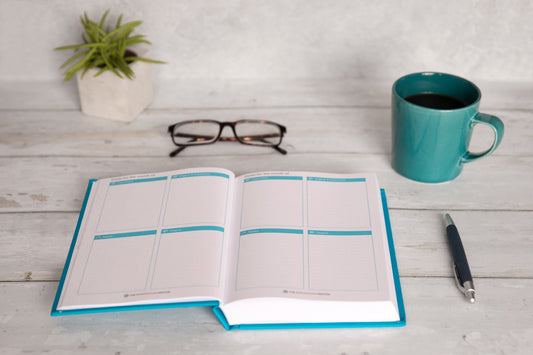 The Empowerment Planner displayed on a desk with a mug, a pen and reading glasses, showing the weekly and daily pages of  this 420-page goal setting journal